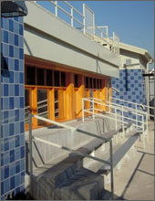 Photo of the exterior of a tennis club. The building has columns on the far right and left side covered with light blue and dark blue tile. Between the columns are three rows of cement bleachers with hand rails. Above the bleachers are windows with wood frames. Above the windows is an open viewing platform with an overhang above the platform. It is implied that these bleachers look out on the tennis court.